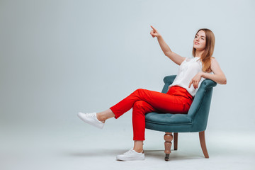 girl in red pants sits in a chair and points at a light gray background. copy space