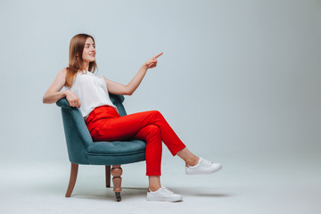 Fototapeta na wymiar girl in red pants sits in a chair and points at a light gray background. copy space