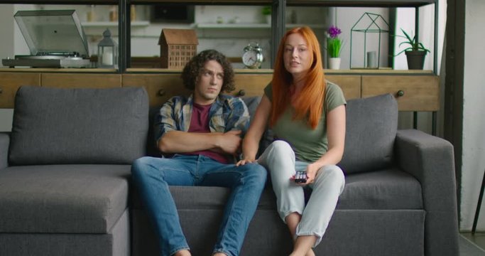 Young couple is sitting on sofa in living room, woman is in good mood, asking man to watch TV with her, man is bored. Caucasian, white. 4K, Middle plan, shot on RED camera.