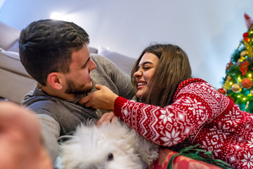 Smiling couple taking selfie with their dog. In background Christmas tree. Christmas holidays concept.