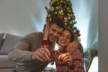Cropped image of happy young couple holding sparklers, hugging and smiling while celebrating Christmas at home
