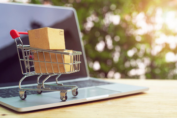 Brown paper boxs in a shopping cart with laptop keyboard on wood table on the public park background.Easy shopping with finger tips for consumers.Online shopping and delivery service concept.