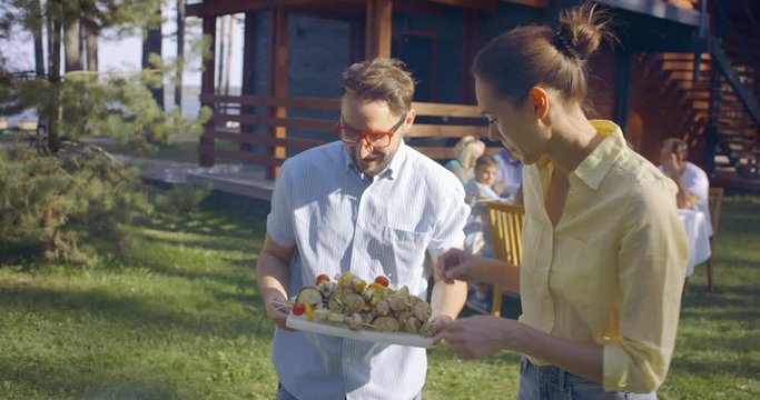 Couple preparing vegetarian barbecue kebabs for large multi generation extended family, people enjoying summertime outdoors. 4K UHD