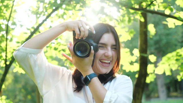 Pretty smiling and laughing girl photographer wearing white shirt is making photos in a park on a soft background of green foliage and spraying water.