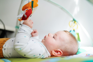 Side view of beautiful curious caucasian six months old baby boy lying in bed and playing with crib toys.