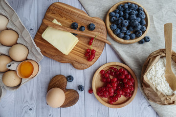 ingredients for baking lie on a light wooden background with a blue kitchen towel. flour, eggs, butter, berries, and wooden tools