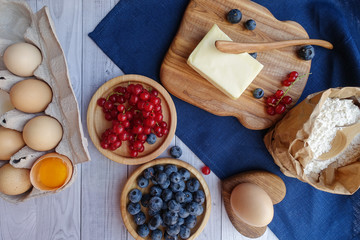 ingredients for baking lie on a light wooden background with a blue kitchen towel. flour, eggs, butter, berries, and wooden tools
