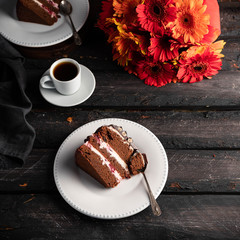 chocolate cake slice on a white plate, Gerber daisy flowers bouquet on a dark wooden backdrop
