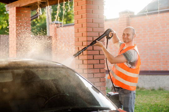 A working man in orange vest washes his car with a large head of water from a karcher on open air. High pressure cleaning
