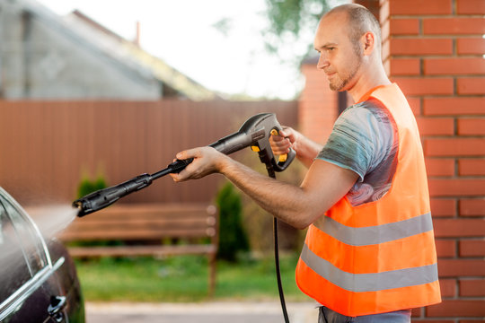 A man in orange vest washes his car with a large head of water from a karcher on open air. High pressure cleaning