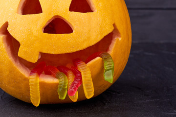 Funny Halloween pumpkin with worms in mouth. Close up carved Halloween pumpkin with jelly woms on dark background. Halloween decoration ideas.