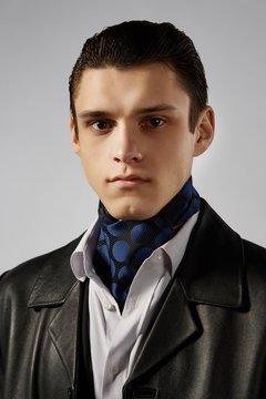 Cropped front view shot of a dark-haired man, wearing white shirt, black jacket and black leather coat. He has black spotted cravat with blue circles print on his neck. Man is looking straight. 