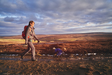 A side view of a hiker and their dog walking along a dirt track that crosses Edmundbyers moor at sunset.