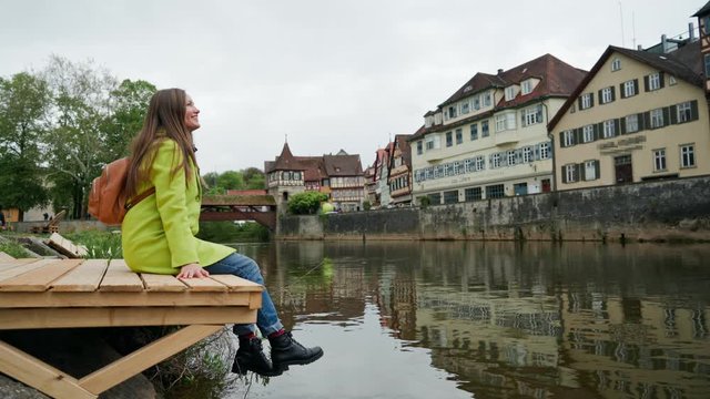 Girl in yellow coat with backpack sitting dangling on a wooden pier near water and looking at traditional german half-timbered houses. Schwabisch Hall. Germany. Side view