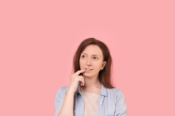 Dreaming young beautiful woman looking up put finger to mouth posing on pink background. Pensive red-haired girl dream of discounts and gifts. Advertising space