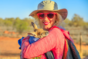 Tourist woman holding kangaroo joey at sunset in Australian outback. Interacting with cute kangaroo...