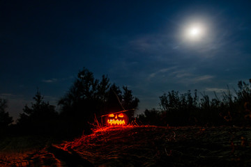 Fototapeta premium Carved halloween pumpkin jack-o-lantern wearing witch hat with burning candles glows in a darkness. Spooky landscape