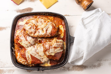 Parmegiana Steak also known as Filet Parmegiana in a black iron pan on a wooden white background, cheese and tomato sauce. Soft light, top view. Flat layer