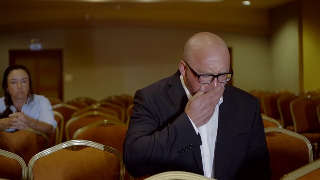 A middle-aged man and woman sit in the audience and watch a lecture on the screen, a man sits in the foreground and occasionally falls asleep, then regains consciousness