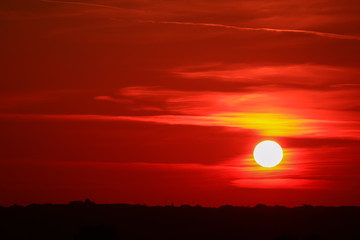 Close up on the sun rising behind a hill on the horizon. Sky with red and orange stratus clouds. Romantic and beautiful sunrise with vegetations of trees.