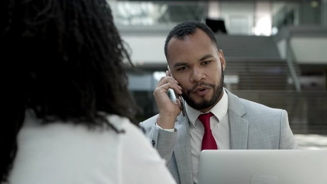 Thoughtful young businessman talking on smartphone. Focused African American man communicating with interlocutor while sitting at table with colleague. Business communication concept