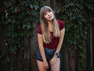 Lifestyle happy blonde woman dressed red blouse blue denim jeans shorts emotionally posing ivy wooden fence european cozy village in evening twilight