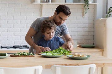 Young father teaching little focused school kid son slicing vegetables.