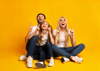 Parents And Daughter Pointing Fingers Upward Sitting Over Yellow Background