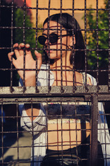 Portrait of girl in sunglasses posing in city behind a trellised fence. Dressed in top with floral print, white shirt, black trousers, waist bag.