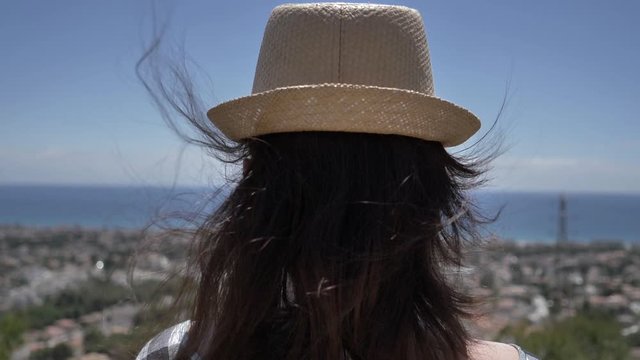 traveler girl in a hat looks down on the city and the sea close up, back view, slow motion