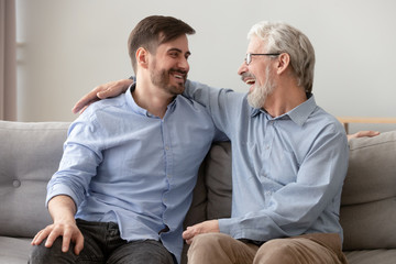 Happy old dad embracing young son talking laughing on sofa