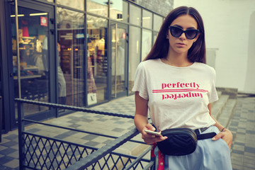 Portrait of a girl in sunglasses posing near a city mall, using smartphone. Dressed in white t-shirt, blue trousers, black waist bag, red kerchief.