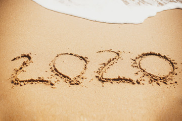 Happy New Year 2020, handwritten on sandy beach. 2020 numbers inscription on golden sand and waves foam at sea beach. Tropical celebration