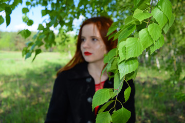 View through the green foliage of a red-haired girl in a coat in nature.