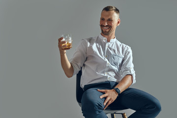 Man with stylish mustache, in classic white shirt and blue trousers is sitting on chair, holding glass of whiskey. Grey background, close-up shot.