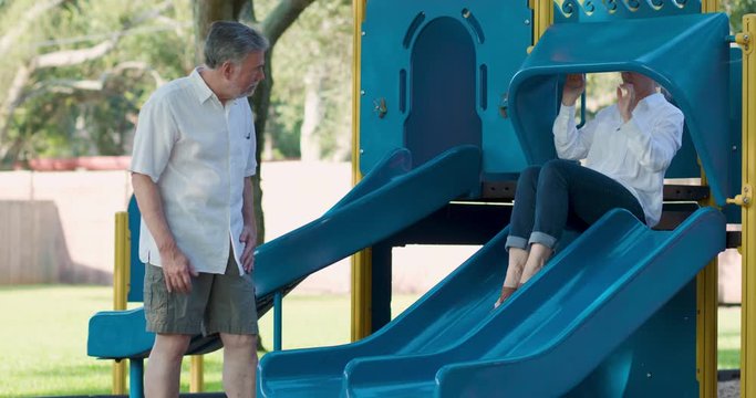 A mature woman playfully sliding down a small playground slide experiences some discomfort in her back.