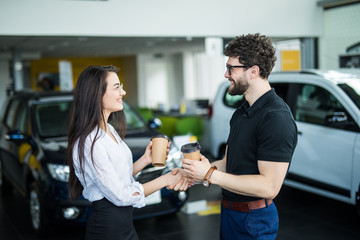 Beautiful girl is drinking coffee near new car. She is in dealership looking for new car. Car is red. She is smiling.