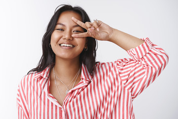 Cheerful young south-asian girl in striped blouse, smiling carefree, showing victory or peace sign...