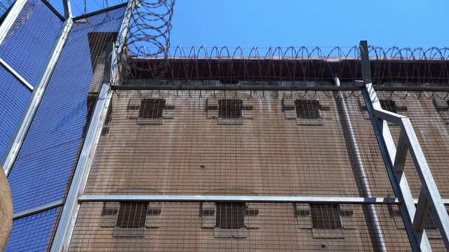 Prison wall with small windows with bars and a high fence with barbed wire