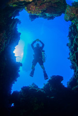 A shot of a scuba diver taken through a window in the coral reef. The silhouette of a dive boat can be seen floating on the surface way above