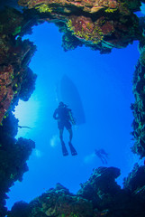 A shot of a scuba diver taken through a window in the coral reef. The silhouette of a dive boat can be seen floating on the surface way above