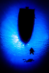 Silhouette shot of divers underneath a boat. The sun can be seen in the sky above the deep blue water. 