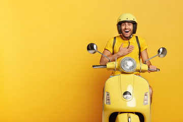 Joyful pleased man receives good news directly on his way, keeps hand on chest, wears helmet, poses on fast moped, isolated over yellow studio wall, blank space aside for your advertisement.