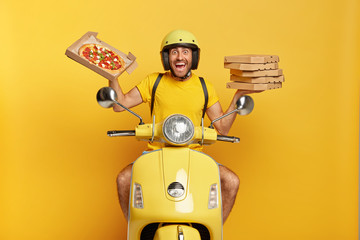 Our most delicious pizza for you. Cheerful young man holds opened pizza box, drives yellow moped, being in hurry to transport fast food in time, wears protective helmet, has much duties at work