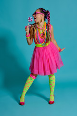 Lovely girl with a multi-colored braids hairstyle and bright make-up, posing in studio against a blue background, holding a lollipop in her hand.