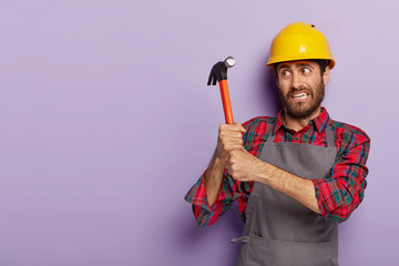 Studio shot of puzzled workman holds hammer, does repairing, wears uniform and protective hardhat,...