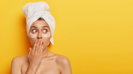 Studio shot of emotional young lady covers mouth with hand, looks with bugged eyes, has bated breath, being topless, looks away, cares about delicate skin under eyes, isolated on yellow studio wall