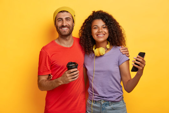 Photo of happy mixed race couple spend free time together, drink coffee and use modern cellular for online communication, dressed in t shirts, stand closely to each other against yellow background