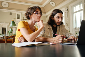 Two young casual students thoughtfully working on study project with laptop in library of university