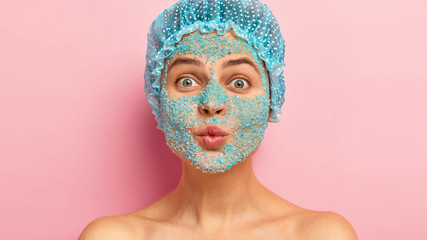 Headshot of beautiful woman with rounded lips, wears white showercap, peels face with organic...